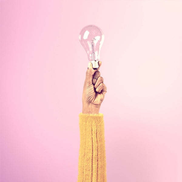 Main levée tenant une ampoule à incandescence allumée, sur un fond rose pastel, évoquant l'inspiration, la créativité et les idées lumineuses.