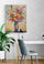 Bureau blanc pieds en bois, chaise grise, objets de décorations tel qu'une lampe, plante, grande plante verte, mur texturé blanc, sol en parquet, affiche murale fleur.