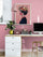 Affiche d'une femme aux fleurs accrochée dans un bureau à la décoration féminine. Le mur est rose, le bureau en bois clair accueille un ordinateur, une plante en pot doré et des boîtes déco. Une suspension rose descend du plafond.