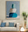 Large modern canvas wall art of three stacked blue and white pebbles, hung above a beige sofa with blue cushions, a designer lamp, and a green plant in a contemporary living room