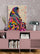 Meuble bas en bois rose pâle au design scandinave, surmonté d’un tableau coloré représentant une femme voilée dans un patchwork géométrique vibrant. Le mur est habillé d’un papier peint à motif floral en spirale. L’ensemble crée une ambiance douce et artistique, ponctuée d’objets déco modernes comme une lampe design arrondie et un vase texturé.