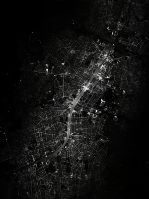 The black and white image shows a nighttime aerial view of illuminated roads winding through the dense urban fabric of a city.