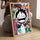 Seated person holding a framed anime print with a smiling character over a vibrant graffiti background; wooden frame on their knees in a bright room