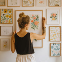 een vrouw op de rug gezien hangt een bloemenschilderij op in het midden van een witte muur versierd met bloemenlijsten. Het tafereel baadt in natuurlijk licht, omringd door groene planten en gevarieerde bloemenmotieven, in een zachte en rustgevende sfeer.