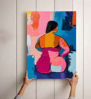 Colorful painting held by two hands, depicting a woman from the back with generous curves, in a knife painting style. The piece blends vibrant patches of pink, orange, blue, and yellow, creating an expressive and modern composition. The textured background with vivid colors evokes contemporary art and the celebration of bodies.