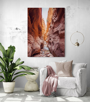 The canyon artwork is hung in a spacious and bright living room. Below, there's a cozy white sofa draped with a pale pink throw. Next to the sofa, a lush green plant adds a nature touch to the room. A minimalist designed hanging light completes the living room vibe.