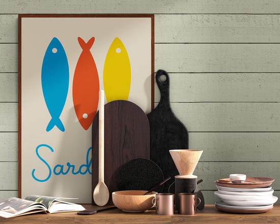 Wall poster depicting three stylized sardines in blue, red, and yellow, resting on a wooden countertop in a rustic kitchen, surrounded by utensils, cutting boards, and dishes.