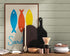 Wall poster depicting three stylized sardines in blue, red, and yellow, resting on a wooden countertop in a rustic kitchen, surrounded by utensils, cutting boards, and dishes.