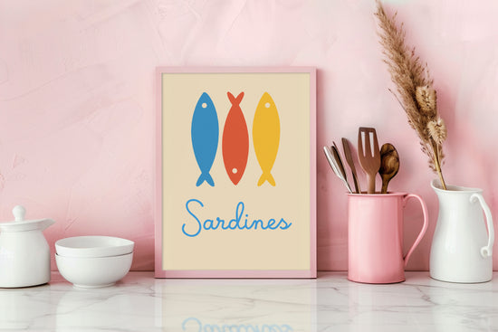 Decorative frame placed on a pink countertop in a modern kitchen, featuring three stylized sardines in blue, red, and yellow with "Sardines" text, bringing a joyful and colorful ambiance.