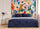 Sophisticated bedroom with a bed dressed in midnight blue linens and a simple headboard. In the background, a large floral tapestry in bright hues of pink, orange, violet, and green. Two golden tables, a white lamp, and a vase of purple flowers create a soft and chic balance.