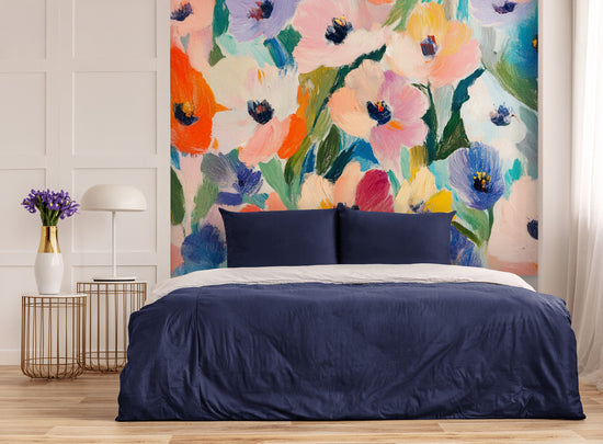Sophisticated bedroom with a bed dressed in midnight blue linens and a simple headboard. In the background, a large floral tapestry in bright hues of pink, orange, violet, and green. Two golden tables, a white lamp, and a vase of purple flowers create a soft and chic balance.