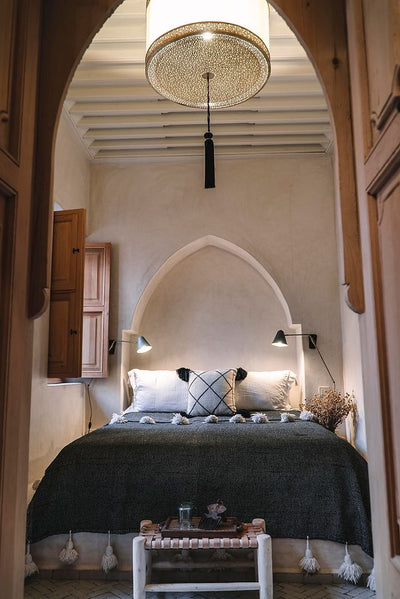 Elegant Moroccan-style room with carved arch headboard, metal lattice ceiling light, Berber pompom cushions, and dark woven cover. The atmosphere blends craftsmanship, comfort, and oriental architectural charm.