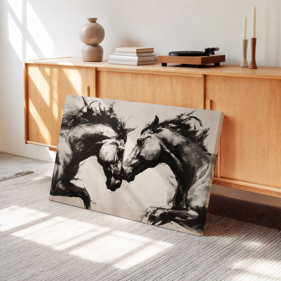 Artwork of two facing black and white horses, placed on the floor in a bright living room with light wooden furniture. Natural light highlights the texture of the canvas and the energy of the equestrian duo, creating a soothing and elegant atmosphere, perfect for natural and authentic decor.