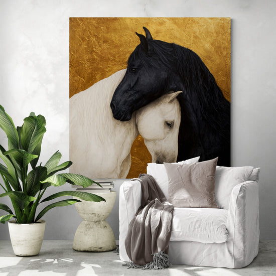 Decorative painting featuring two entwined horses, one black and one white, on a textured gold background, hanging in a modern living room with a white armchair, gray cushions, and a green plant.