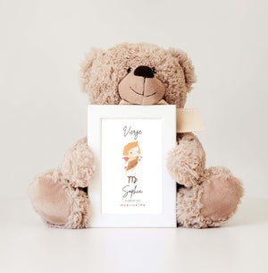 A teddy bear holds a white frame displaying an illustration of the Virgo astrological sign. The image features a small angel with curly hair, a beige tunic, and small pink wings, gently floating surrounded by stars. The name "Sophia" and the birth details are inscribed at the bottom, adding a personalized touch. The background is clean and bright, highlighting the scene's delicacy.