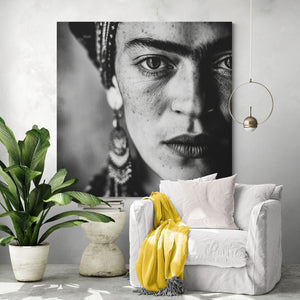Affichage mural moderne avec icône mexicaine: Un grand tirage de Frida Kahlo habille un mur clair. Le fauteuil blanc contraste avec un plaid jaune vif. À ses côtés, une plante verte, une table en pierre, et une suspension dorée. L’ambiance mêle force et douceur.