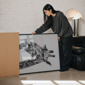 A person holding a large painting of a gently touching giraffe and calf, with the painting placed on the floor near a box in a bright living room.