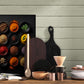 Un tableau de diverses épices colorées dans des bols noirs est accroché sur un mur en bois peint en vert pâle, à côté d'ustensiles de cuisine en bois, d'une cafetière et de vaisselle empilée, créant une ambiance rustique et accueillante.