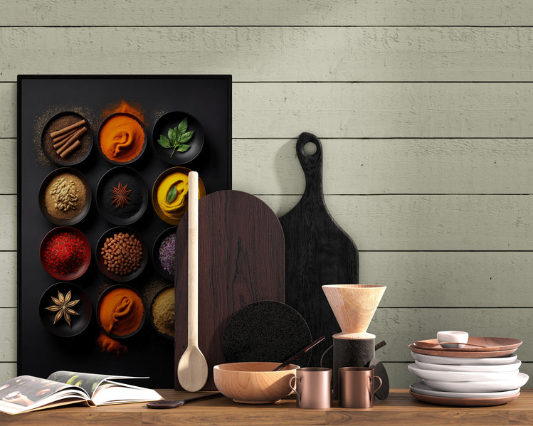 Un tableau de diverses épices colorées dans des bols noirs est accroché sur un mur en bois peint en vert pâle, à côté d'ustensiles de cuisine en bois, d'une cafetière et de vaisselle empilée, créant une ambiance rustique et accueillante.