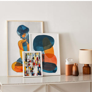 Three framed artistic wall posters on a living room cabinet. The first depicts a stylized female silhouette in blue and orange, the second showcases overlapping abstract shapes in blue, orange, and ochre, and the third features a colorful geometric stained glass-like composition. On the right, a beige lamp and two ceramic vases complete the modern and warm interior decoration.