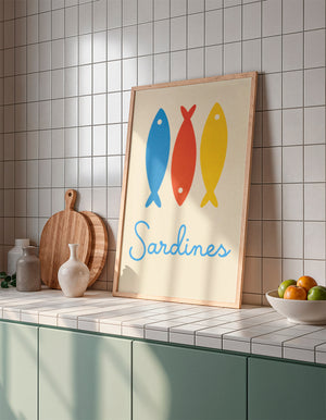 Tableau illustré de trois sardines colorées bleu, rouge et jaune avec l’inscription "Sardines", exposé dans une cuisine carrelée au style méditerranéen, à côté de planches et d’un bol de fruits.