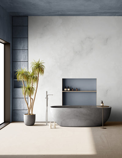 Salle de bain épurée avec une baignoire en pierre grise, une grande plante verte en pot, des murs aux tons neutres et un plafond bleu texturé. Une niche murale bleue accueille des flacons de soins. L’ensemble crée une ambiance zen, naturelle et apaisante, idéale pour la détente