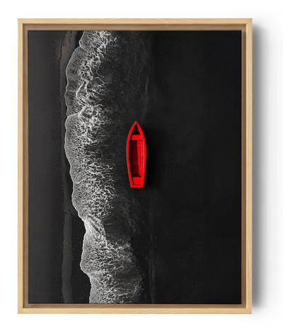Vue aérienne d’une barque rouge flottant sur une mer noire, près d’une plage au sable sombre, avec des vagues blanches créant un contraste saisissant.