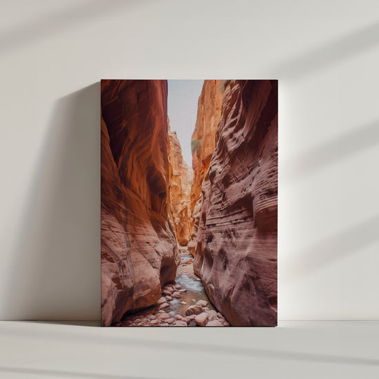 Narrow red sandstone slot canyon carved by erosion with a pebble-strewn stream on the floor and blue sky above