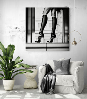 Contemporary interior with a white armchair, a dark throw, and a black and white canvas depicting women's legs in high heels and thigh-high socks. The image plays on urban elegance and graphic contrast, creating a chic and bold ambiance