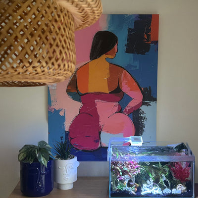 Customer photo featuring a colorful contemporary painting of a curvy woman seen from behind, styled in a modern interior with a woven lamp, two plants, and an aquarium on a console table.