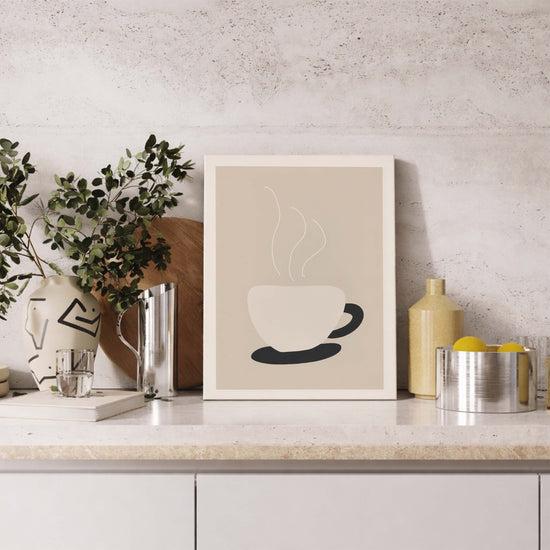 Affiche minimaliste représentant une tasse de café blanche fumante, imprimée sur toile canvas et posée sur un plan de travail de cuisine moderne. Le tableau aux tons beige et noir s’intègre parfaitement dans une décoration épurée, avec mur en béton clair, vase décoratif, feuillage vert et accessoires en métal argenté. Une œuvre murale idéale pour sublimer une cuisine contemporaine avec une ambiance chaleureuse et naturelle