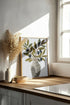 Art print depicting a striped vase filled with olive branches and black olives, sitting on a wooden countertop, bathed in natural light from a window. The scene exudes a soft, Mediterranean ambiance.