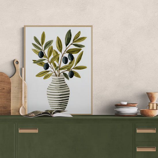 A decorative print featuring a striped black and white vase filled with olive branches and black olives, placed on a dark green kitchen cabinet. Surrounding elements include wooden boards, dinnerware, and a manual coffee maker, creating a warm, Mediterranean atmosphere. The scene radiates simple, natural elegance.