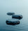 Un papier peint panoramique représentant trois galets posés sur une eau calme et paisible, avec une brume douce à l'arrière-plan. Les couleurs bleutées et apaisantes créent une atmosphère zen