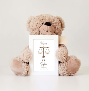 White frame with an illustration of the Libra astrological sign, held by a plush teddy bear against a neutral background.