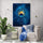 A modern canvas featuring a peacock feather painted in deep blue, with golden hues in the center. The frame is hung above a white upholstered armchair, adorned with a light blue cushion and throw. The space design emphasizes simplicity and elegance.