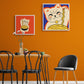 Dans une salle a manger les murs sont orange au centre une table et des chaise au style industrielle. deux tableaux de chat maneki-neko sont accroché au mur
