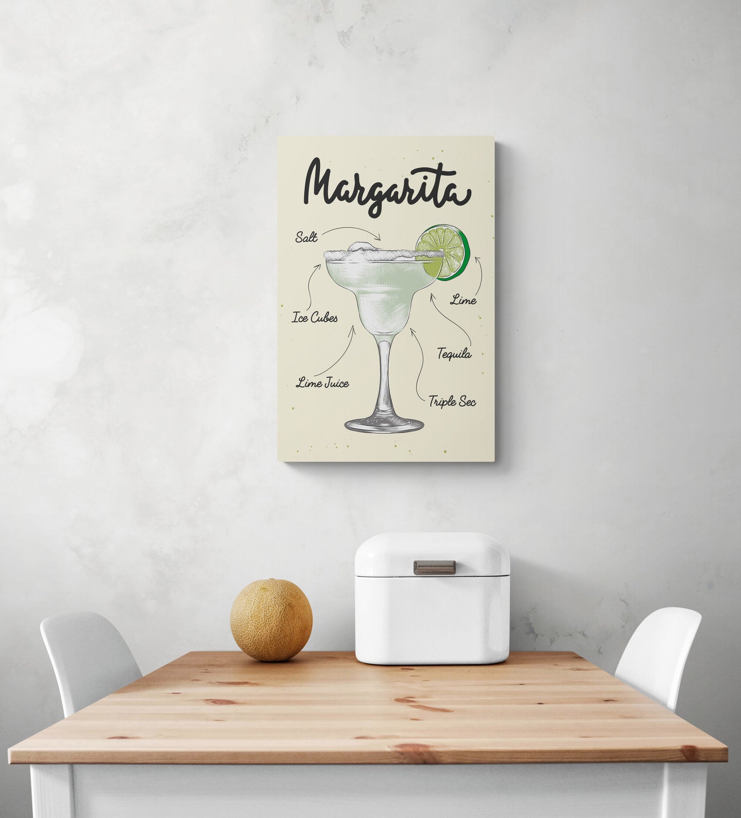 Un cadre deco cuisine jaune avec une illustration d'un verre de cocktail Margarita et les ingrédients écrits en typographie manuscrite sur un fond blanc. Une table à manger en bois et deux chaises blanches se trouvent sous le cadre, et une corbeille à pain et un melon se trouvent au-dessus.
