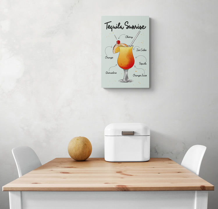 Un cadre cuisine est place au centre d'un mur blanc. Le petit tableau déco pour cuisine montre un verre de cocktail de tequila. L'intérieur est décoré de façon minimaliste avec deux chaises blanches à chaque extrémité. Une boîte à pain en métal blanc et un fruit sont posés sur la table.