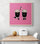 A pink decorative art piece hung on a white kitchen wall above a wooden table flanked by two chairs. A white metal bread box and a melon sit on the table.