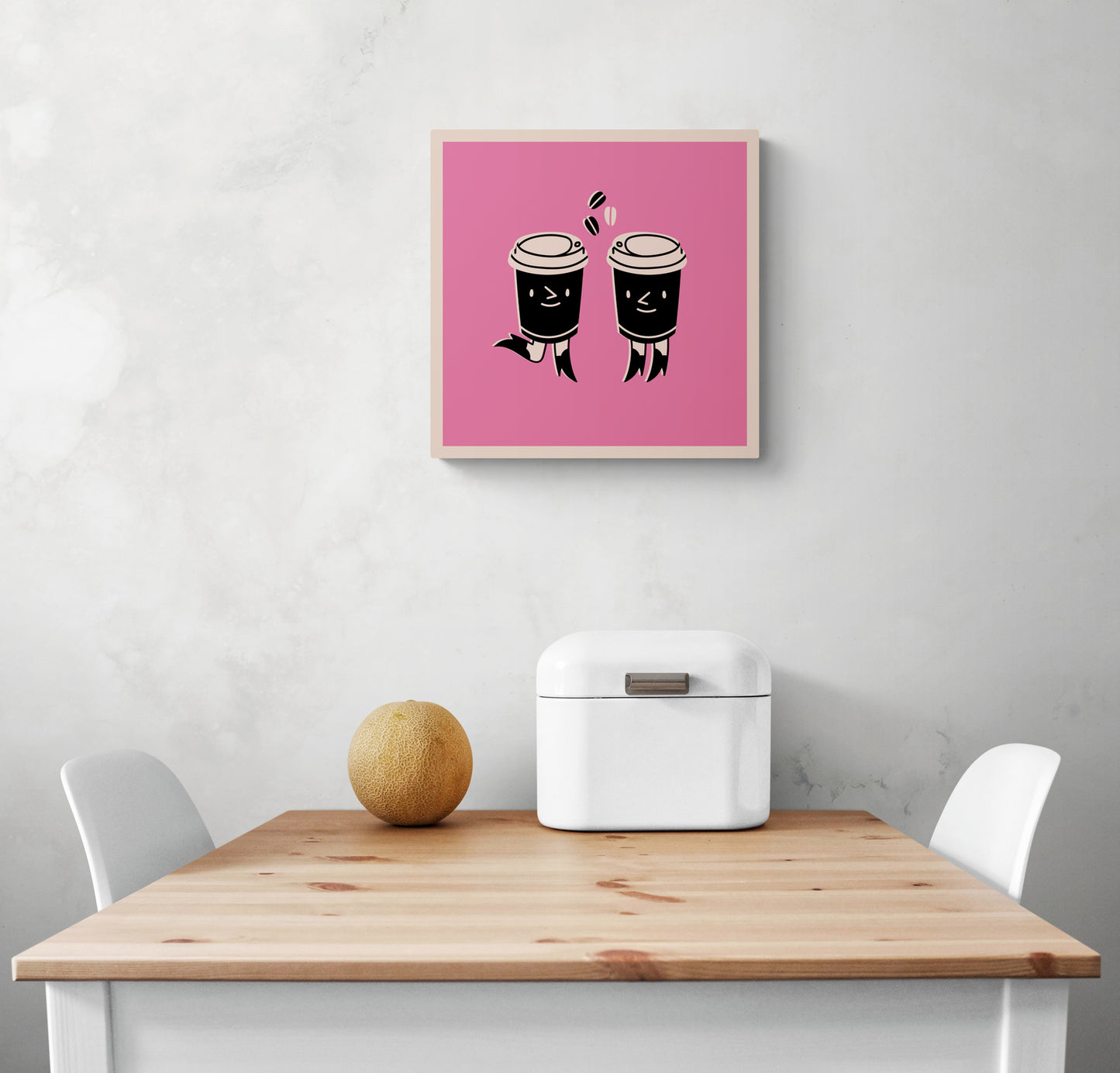 Un petit Tableau rose dans une cuisine blanche. Ce tableau cuisine est placé au-dessus d'une table en bois et deux chaises sont de chaque côté. Une boîte à pain en métal blanc et un melon sont sur la table
