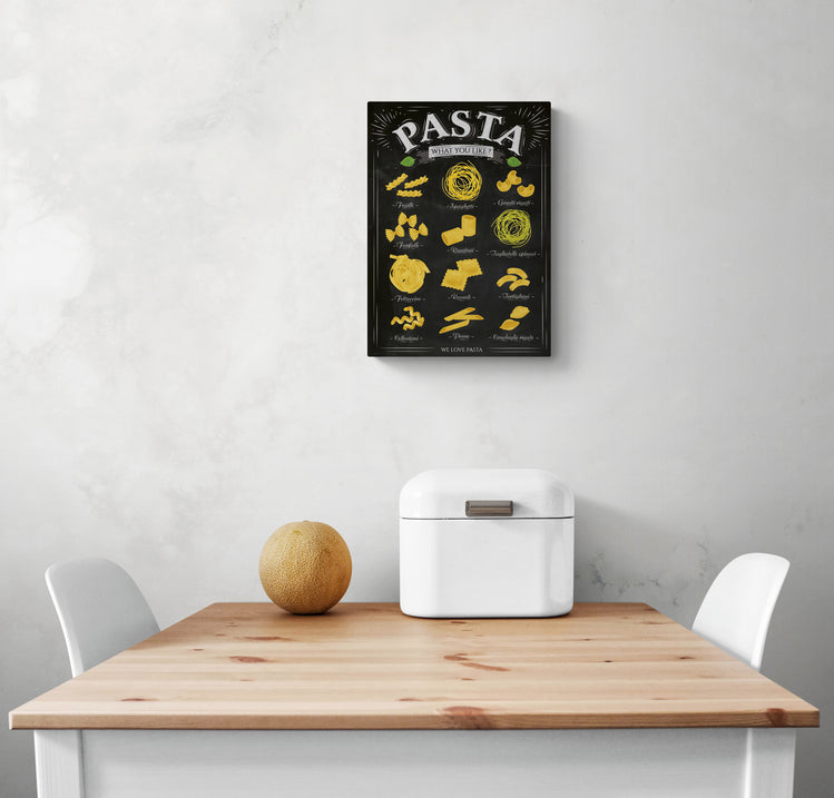 Sur un mur de cuisine blanc, un tableau noir encadré avec différents types de pâtes est positionné au centre. Sur la partie inférieure du mur, une table à manger en bois et deux chaises blanches sont présentes. Une corbeille à pain en métal blanc et un melon sont positionnés sur la table.