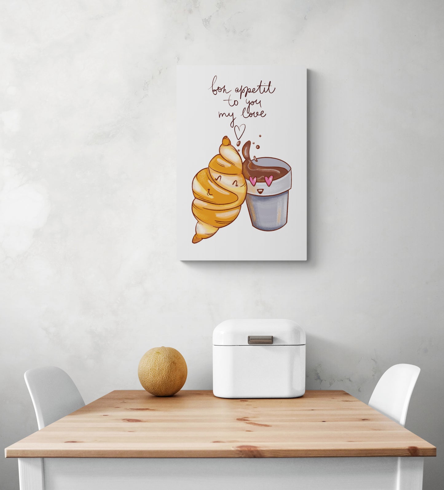 Un tableau petit format pour cuisine est accroché au milieu d'un mur blanc. Ce tableau cuisine est placé au-dessus d'une table en bois et deux chaises sont de chaque côté. Une boîte à pain blanche et un melon sont posés sur la table