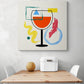 Un tableau pour cuisine moderne sobre et épuré avec l'illustration d'un verre de vin autour des formes géométriques et des formes aux pinceaux orange, vert, bleu, jaune, rouge. Ce tableau cuisine est placé ce au-dessus d'une table en bois et deux chaises sont de chaque côté.