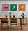 Dans une salle à mangé épuré au ton gris trois cadres colorés, orange, rose, et vert sont accrochés sur le mur. Une table et deux chaises en bois sont sur l'image