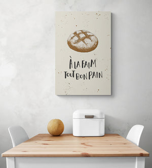 Un tableau beige avec une illustration de pain de campagne est suspendu au mur blanc. Sous le tableau cuisine une table et deux chaises blanches