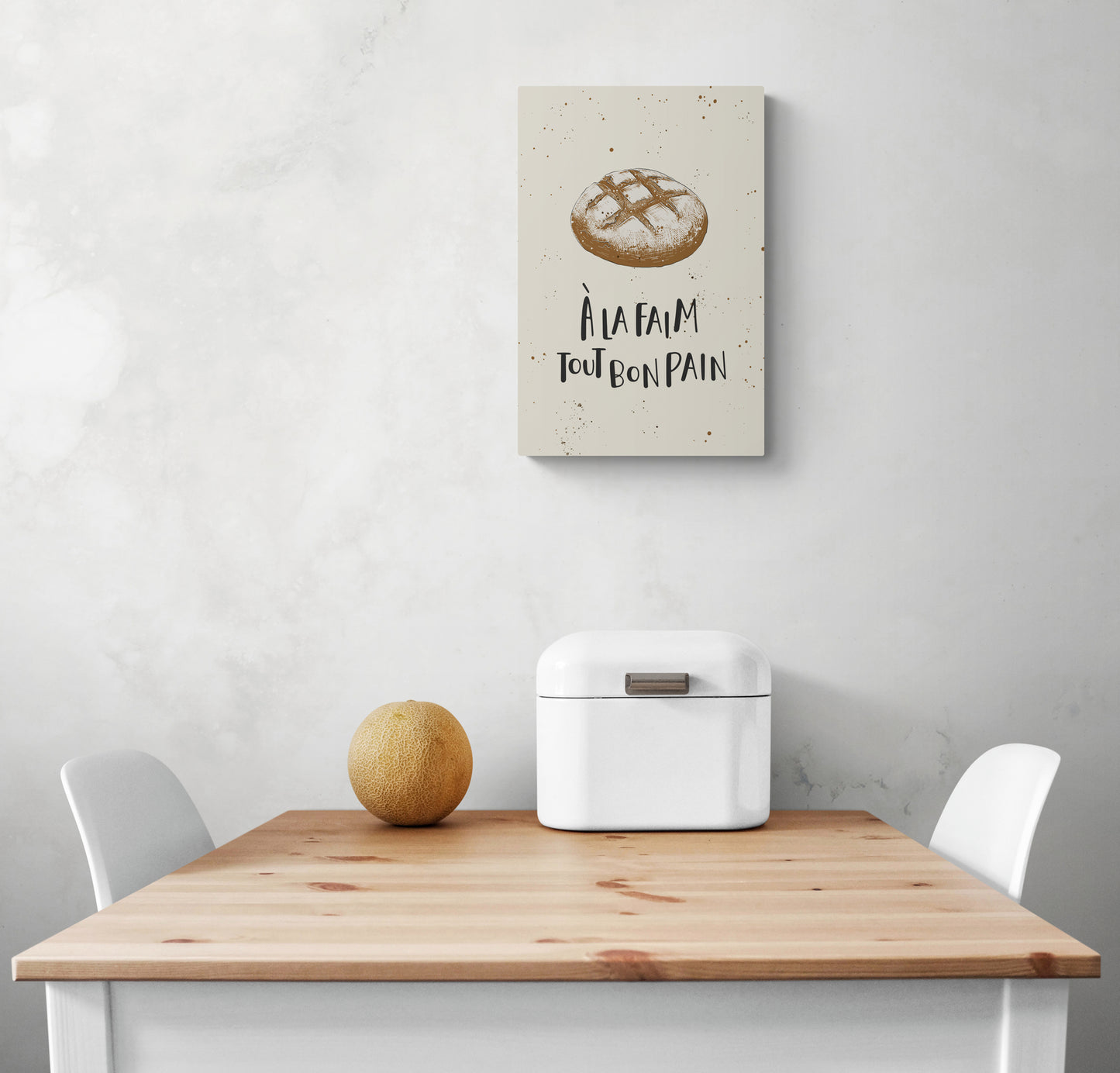 Un petit tableau de décoration beige avec une illustration de pain de campagne est suspendu au mur blanc. Sous le tableau cuisine une table et deux chaises blanches 