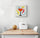 Modern kitchen wall art centered on a white wall; a minimalist canvas with a black wine glass illustration surrounded by geometric shapes—square, rectangle, triangle, and circle—and painterly brush marks; below, a white dining table with a white chair at each end, plus a melon and a white bread box on the tabletop