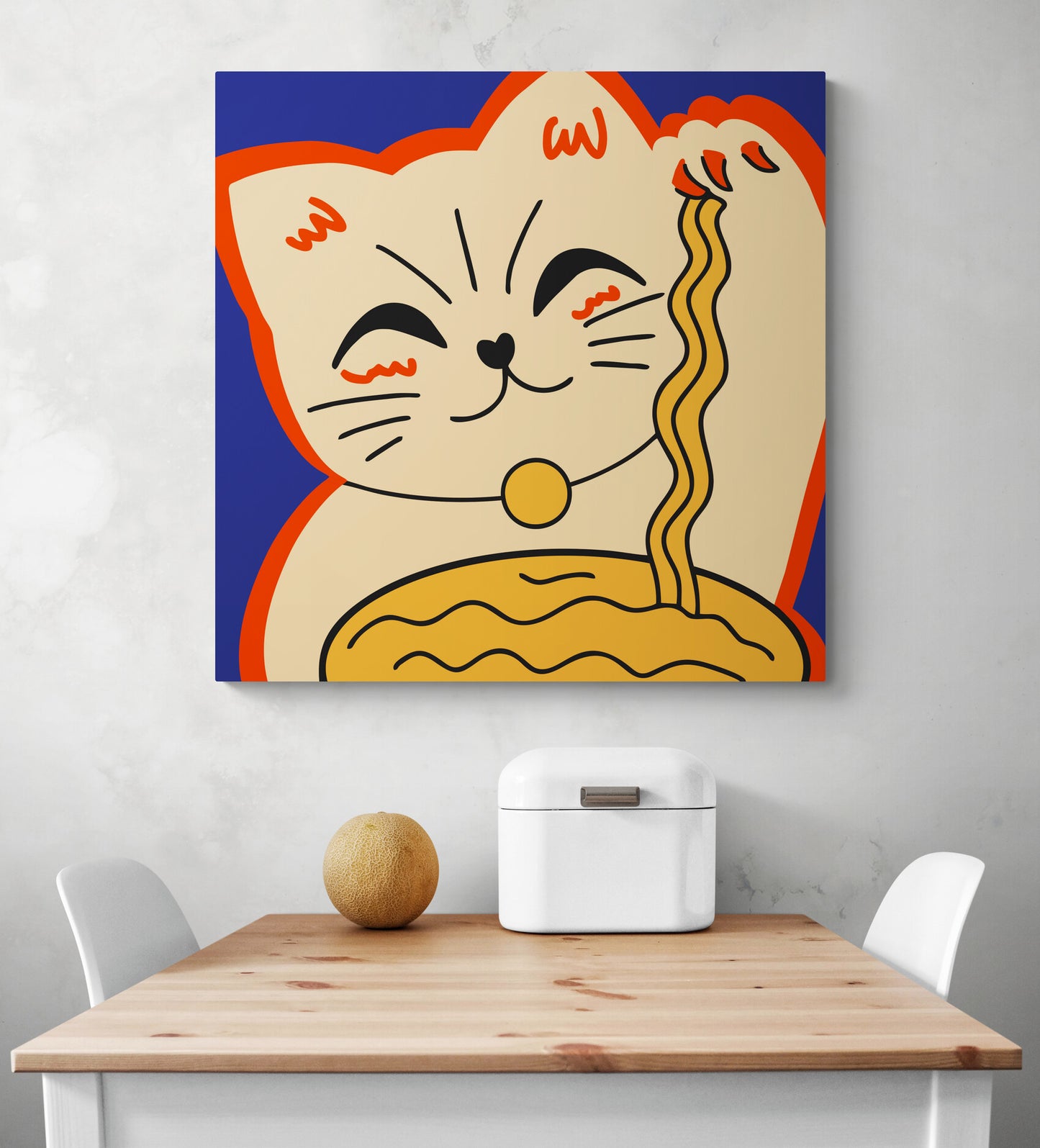 Dans une cuisine blanche sur un pan de mur est suspendu un tableau de décoration bleu, au centre du tableau déco une illustration d'un chat Maneki-neko. Il tient dans ça patte droite des nouilles chinoise. En dessous une table de cuisine en bois et deux chaises blanches