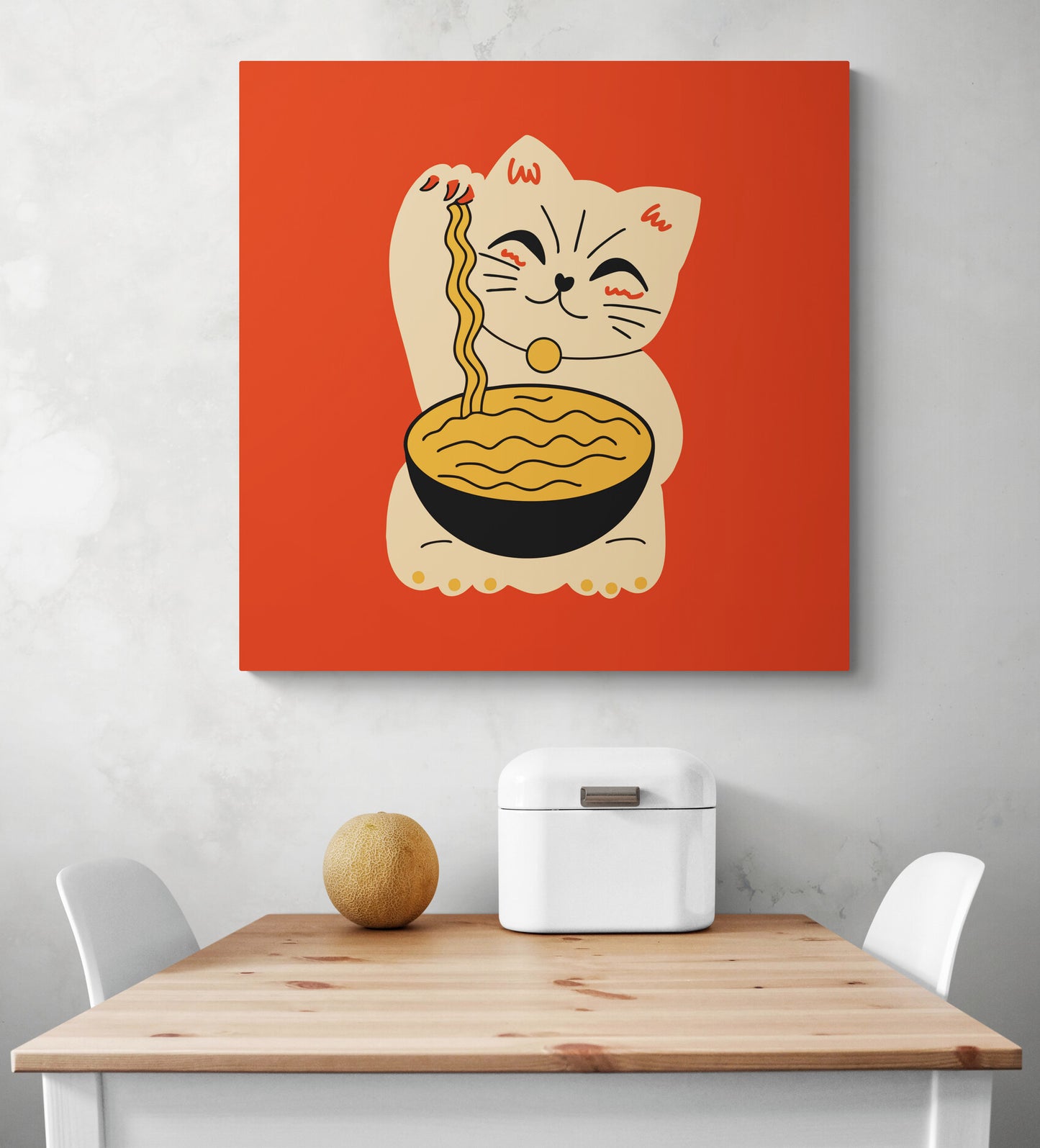 Dans une cuisine blanche sur un pan de mur est suspendu un tableau de décoration orange, au centre du tableau déco une illustration d'un chat Maneki-neko. il tient dans ses pattes un bol noir de nouille. en dessous une table et deux chaises blanches